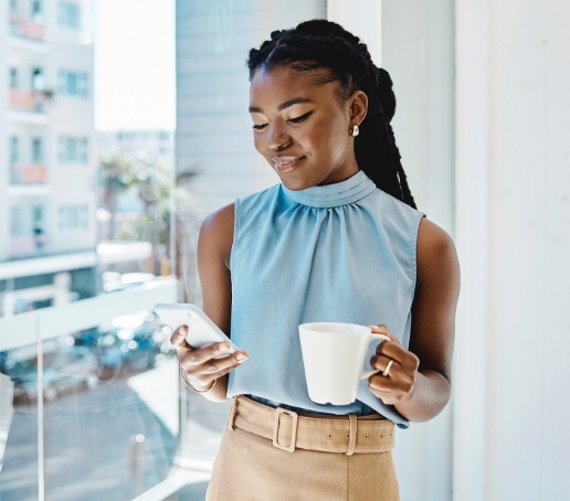 Businesswoman in the office looking at her cellphone and holding a cup of tea