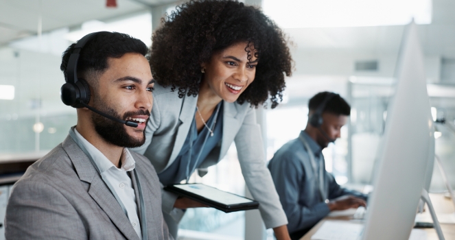 Business man in an office with a headset viewing a computer monitor with a business woman looking over his shoulder. Both are smiling.